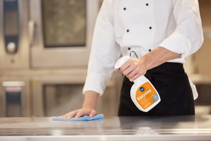 Kock desinfekterar rostfri arbetsyta i restaurangkök med Kiilto Pro Easydes Spray och blå mikrofiberduk.