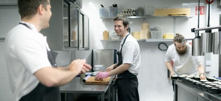 Tre kockar arbetar i ett professionellt restaurangkök med rostfria arbetsbänkar.